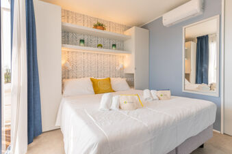Double bed with white bedding and a yellow pillow under shelves with decorative items and a large mirror at Le Mimose Family Resort