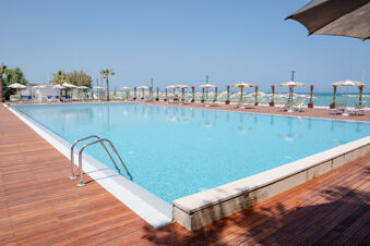 Outdoor swimming pool with wooden deck, sun loungers, umbrellas, palm trees, and a view of the sea at Le Mimose Family Resort