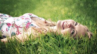 A woman wearing a floral dress is lying on green grass smiling outdoors with Pension Graml Fr&uuml;hst&uuml;ckspension branding visible