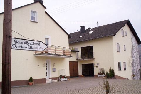 Two connected light-colored buildings with balconies and a sign for Hotel - Pension Wendland on a wooden pole in front of them