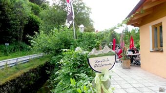 V&auml;liala koos tervituskilbiga ja k&uuml;lalised laudade juures hotelli Restaurant Pension Weiherm&uuml;hle terrassil rohelises, looduslikus keskkonnas.