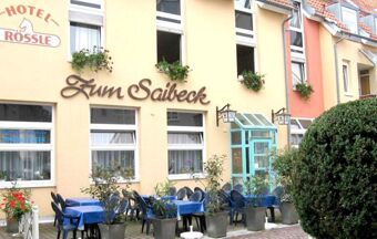 Au&szlig;enansicht des Hotels R&ouml;ssle mit einem Restaurantbereich "Zum Saibeck" und gedeckten blauen Tischen im Freien vor gelben und orangefarbenen Hausfassaden.