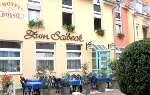 Au&szlig;enansicht des Hotels R&ouml;ssle mit einem Restaurantbereich "Zum Saibeck" und gedeckten blauen Tischen im Freien vor gelben und orangefarbenen Hausfassaden.