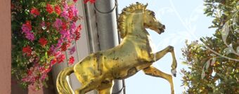 Nahaufnahme einer goldenen Pferdeskulptur an der Fassade mit roten Blumen und B&auml;umen, typisches Detail vom Hotel R&ouml;ssle.