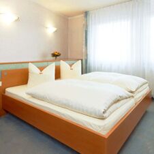 A neatly made double bed with white linens and pillows in a bright room at Hotel Hahnen with a window and wall lamps