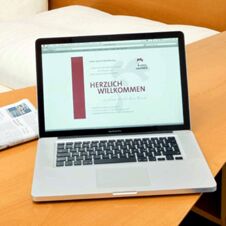 Laptop med Hotel Hahnen-velkomstskjermen &aring;pen p&aring; et trebord ved siden av en seng p&aring; et hotellrom.