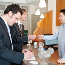 Two men checking in at Hotel Hahnen reception desk with a receptionist handing over a keycard in a bright room
