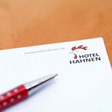 N&aelig;rbilde av en notisblokk fra Hotel Hahnen med logo og kulepenner p&aring; et trebord, egnet for notater og informasjon.