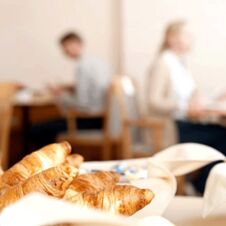 N&aelig;rbilde av ferske croissanter p&aring; et bord i frokostomr&aring;det p&aring; Hotel Hahnen, med gjester i bakgrunnen.