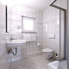 Bathroom in Hotel Hahnen with white tiled walls and floor, wall-mounted sink, toilet, towel rack, and shower area visible