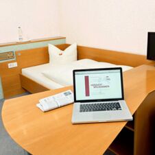 A single bed with white linens next to a wooden desk holding an open laptop displaying Hotel Hahnen&rsquo;s welcome page and a folded newspaper