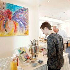 A woman and a man serve themselves drinks from a buffet table in a bright room at Hotel Hahnen with a colorful rooster painting on the wall