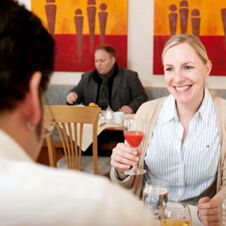 A smiling woman holding a glass of red drink sitting at a table in the dining area of Hotel Hahnen
