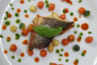 A piece of cooked fish topped with a green basil leaf, surrounded by small carrots, potatoes, and green and red sauce dots on a white plate from Winzerhotel Weingut Kinges-Kessel