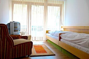 A bright room at Landgasthof Hirsch Dapfen with two single beds, a striped armchair, a small TV, and sheer curtains over a balcony door