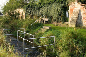 Naturnahe Au&szlig;enansicht mit einer Metallbr&uuml;cke &uuml;ber einen Bach und einer alten Mauer als Kulisse beim Hotel Restaurant Klosterhof GmbH.
