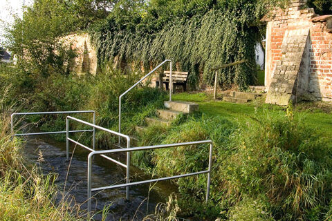 Naturnahe Au&szlig;enansicht mit einer Metallbr&uuml;cke &uuml;ber einen Bach und einer alten Mauer als Kulisse beim Hotel Restaurant Klosterhof GmbH.