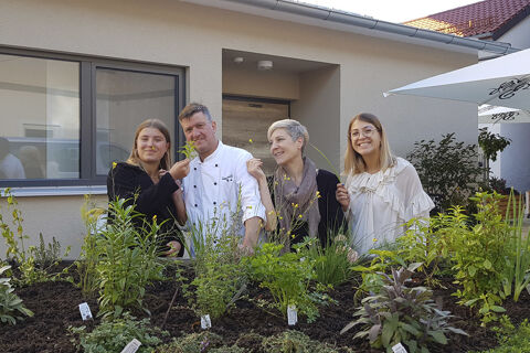 Vier Personen, darunter ein Koch, sitzen lachend hinter einem Kr&auml;utergarten vor dem Geb&auml;ude des Hotel Restaurant Klosterhof GmbH.