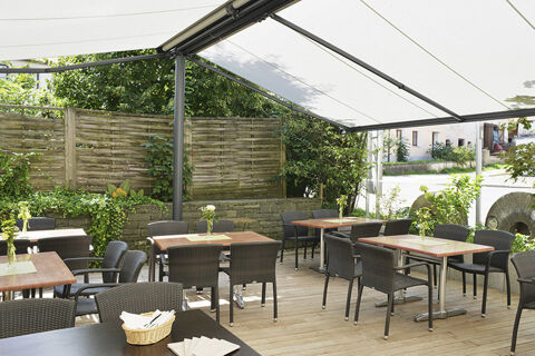 Sonnige Terrasse mit modernen Tischen und St&uuml;hlen im Au&szlig;enbereich des Hotel Restaurant Klosterhof GmbH, umgeben von gr&uuml;nem Bewuchs.
