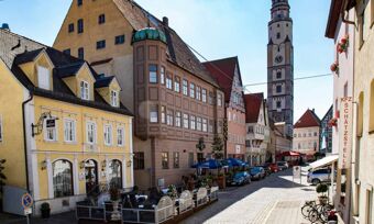 A sunny cobblestone street with parked cars, bicycles, shaded outdoor seating, and the Lodner&acute;s Genie&szlig;erhotel & Hotel Drei Mohren buildings