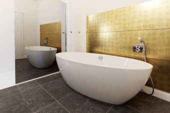 Modern white freestanding bathtub with gold tiled wall and dark patterned floor in Lodner&acute;s Genie&szlig;erhotel & Hotel Drei Mohren bathroom