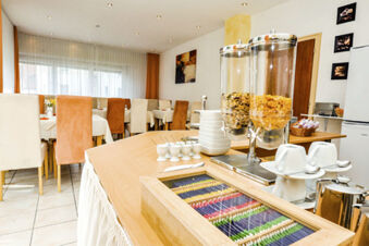 Sala colazione dell'Hotel Garni Metzingen con tavoli apparecchiati, accessori per la colazione e distributori di muesli in un ambiente luminoso.