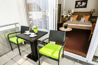 Balcone con tavolo e due sedie con cuscini verdi davanti a una camera con letto matrimoniale dell'Hotel Garni Metzingen.