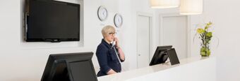 Receptionist talking on the phone at the white front desk with two computer monitors and wall clocks at NH Hotel Frankfurt Messe