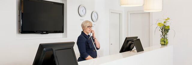 Receptionist talking on the phone at the white front desk with two computer monitors and wall clocks at NH Hotel Frankfurt Messe
