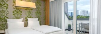 Twin beds with white linens and pillows in a room at NH Hotel Frankfurt Messe with green patterned wallpaper and a balcony view