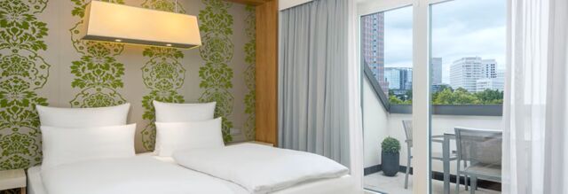 Twin beds with white linens and pillows in a room at NH Hotel Frankfurt Messe with green patterned wallpaper and a balcony view