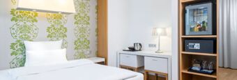 White bed with patterned green wallpaper, a desk with a lamp and electric kettle, and minibar in NH Hotel Frankfurt Messe room