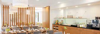 Bright dining area with wooden tables, chairs, decorative plants, buffet with cereals and beverages at NH Hotel Frankfurt Messe