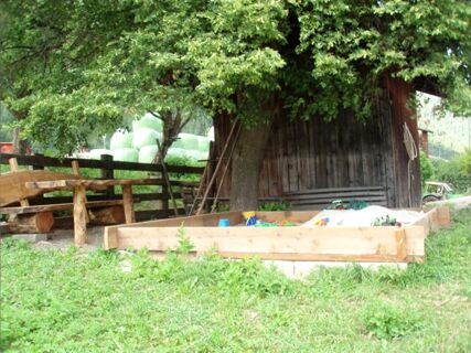 Голям пясъчник с играчки под дърво до дървена къщичка на територията на ваканционните апартаменти на фермата Lexmairhof.
