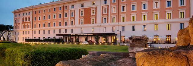 NH Collection Roma Palazzo Cinquecento building illuminated at dusk with green lawn and ancient stone ruins in front