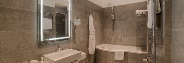 Bathroom with white sink, illuminated mirror, bathrobe, bathtub with showerhead, and towels at NH Collection Roma Palazzo Cinquecento