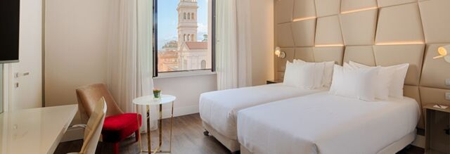 A bright hotel room at NH Collection Roma Palazzo Cinquecento with two white beds, a red chair, small table, and a window view of a bell tower