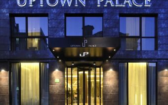 Facade of Uptown Palace Hotel showing dark stone exterior, large glass windows with curtains, and illuminated entrance with revolving door