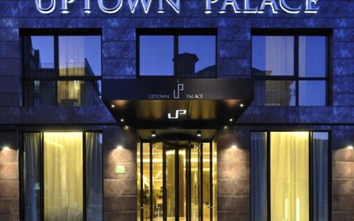 Facade of Uptown Palace Hotel showing dark stone exterior, large glass windows with curtains, and illuminated entrance with revolving door