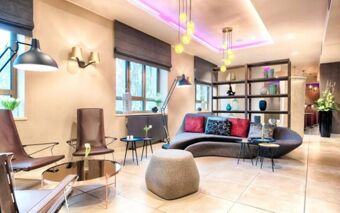 Interior of a modern lounge area at NYX Hotel Milan featuring brown chairs, a curved sofa, glass tables, and decorative shelves