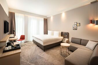 Hell eingerichtetes Hotelzimmer mit gro&szlig;em Bett, Couch, Schreibtisch und Fernseher im Holiday Inn Mannheim City - Hauptbahnhof.