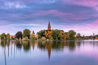Lake view with trees, a tall church steeple, colorful sky, and reflections near Hotel zur Sonne