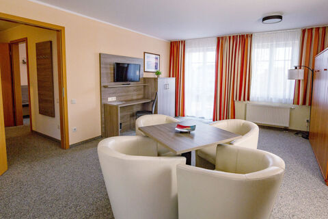 Living area of Hotel zur Sonne with a square wooden table surrounded by four white chairs and a wall-mounted TV above a desk