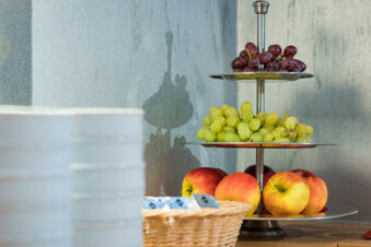 Three-tiered fruit stand holding grapes and apples next to a basket with packets and stacked plates at Hotel zur Sonne
