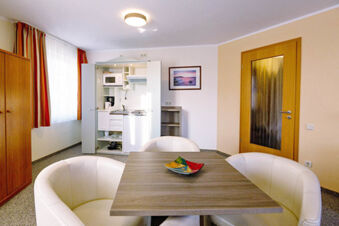 Living area with a small kitchen cabinet, dining table, white chairs, wooden door, and window with orange curtains in Hotel zur Sonne