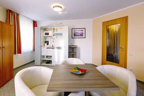 Living area with a small kitchen cabinet, dining table, white chairs, wooden door, and window with orange curtains in Hotel zur Sonne