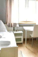 Helles G&auml;stezimmer im G&auml;stehaus Odin mit Einzelbett, Nachttisch, Schreibtisch und Stuhl vor einem gro&szlig;en Fenster mit Vorh&auml;ngen.