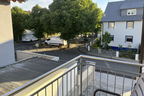 Blick vom Balkon der G&auml;stehaus Odin auf eine ruhige Stra&szlig;e mit B&auml;umen und einem wei&szlig;en Haus mit blauen Fensterrahmen bei Sonnenlicht.