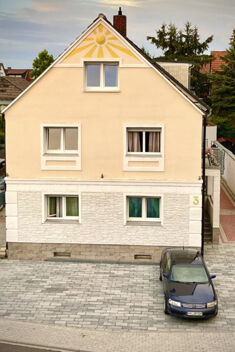 Die Fassade vom G&auml;stehaus Odin zeigt ein zweist&ouml;ckiges Haus mit einem Sonnenmotiv im Giebel und einem geparkten Auto davor.