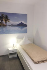 Schlicht eingerichtetes Einzelzimmer im G&auml;stehaus Odin mit wei&szlig;em Bett, Nachttisch und einer Lampe, dazu ein gro&szlig;es Landschaftsbild an der Wand.